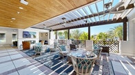 Located in Orlando, FL, this single-level amenity space blends coastal elegance with resort-style comfort. The design draws on a bright, nautical palette of crisp whites, navy blues, and natural wood tones throughout, creating a cohesive and welcoming atmosphere.

The centerpiece of the space is a generous covered outdoor terrace, partially open to the sky via a pergola-style structure with louvered slats that allow natural light to filter through. Ceiling fans keep the air moving, while views of swaying palm trees and the surrounding community contribute to a lush, tropical ambiance. The terrace is furnished with white rattan and wicker chairs accented by vibrant floral and botanical cushions, arranged atop richly patterned area rugs.

Transitioning indoors, the interior lounge area features soaring vaulted ceilings with exposed white-painted beams, recessed lighting, and an eye-catching fireplace topped by a large display screen. Black-framed French doors connect the indoor and outdoor spaces seamlessly, flooding the interior with natural light. Comfortable striped sectional seating and lattice-back chairs are complemented by tropical potted plants, reinforcing the resort feel.

A more intimate lounge nook offers paired light-blue sofas beneath striking wire pendant lanterns, providing a quieter retreat. The tile and hardwood flooring grounds each zone with warmth and texture, tying together this thoughtfully designed communal destination.