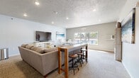Located at 218 19th St SE in Watertown, South Dakota, this single-floor living room offers a thoughtfully organized and versatile communal space. The room is finished with neutral gray carpet and light gray walls, creating a clean and welcoming atmosphere throughout.

The space is divided into three functional zones that flow seamlessly into one another. A comfortable lounge area anchors one side of the room, featuring a plush sofa, tufted ottoman, and a media console supporting a large flat-screen television. Recessed lighting and a textured ceiling add character overhead.

At the center, a rustic wooden bar-height table with industrial-style stools serves as a casual gathering point. Adjacent to this, a bright dining nook is defined by a large multi-pane window that allows generous natural light to pour in and offers views of the surrounding outdoor area. A white round dining table with matching chairs and a bench provides a charming, cottage-inspired setting beneath a bold ocean-themed artwork.

Along the far wall, a freestanding bookcase displays board games and accessories, complementing the room's recreational purpose. A wall-mounted clock and additional coastal artwork reinforce the space's cohesive, relaxed aesthetic. The overall design balances comfort and function, making it ideal for socializing, dining, and leisure activities.