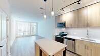 Located at 2311 Dulles Station Blvd in Herndon, Virginia, this modern single-floor residence offers a thoughtfully designed open-concept layout with clean lines and a bright, contemporary aesthetic throughout. Light-toned wood-style flooring flows seamlessly across the living and kitchen areas, creating a cohesive and welcoming atmosphere.

The kitchen is a standout feature, outfitted with light wood cabinetry extending to the ceiling for ample storage, white quartz countertops, a subway tile backsplash, and a full suite of stainless steel appliances including a refrigerator, electric range, built-in microwave, and dishwasher. A central kitchen island with a white countertop and lower storage provides additional prep space and casual seating potential. Elegant pendant lighting hangs above the island, complemented by a track lighting system overhead.

The open living area benefits from expansive floor-to-ceiling windows and corner glazing, allowing abundant natural light to fill the space and offering views of the surrounding neighborhood. The bedroom features the same warm wood-style flooring and generous proportions, enhanced by a large walk-in closet with built-in wire shelving and hanging rods, providing well-organized storage. The overall design emphasizes a clean, modern sensibility with a neutral palette suitable for a variety of interior styles.