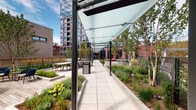 Located in Chicago's vibrant West Loop neighborhood, this outdoor amenity terrace at 22 N Sangamon Street offers a thoughtfully designed urban retreat surrounded by striking contemporary architecture. The space is defined by a sleek, glass-canopied walkway supported by slender steel columns, creating a covered pedestrian corridor that connects the various zones of the terrace while allowing natural light to filter through overhead.

Lush, professionally landscaped planting beds line both sides of the walkway, featuring a diverse mix of ornamental grasses, perennial shrubs, birch trees, and native groundcovers that provide year-round texture and greenery. The hardscaping transitions seamlessly between large-format concrete pavers and warm wood decking, lending visual variety and a refined aesthetic throughout.

A central wood-decked seating area serves as a social hub, anchored by a substantial bowl-shaped fire feature and surrounded by contemporary outdoor chairs — ideal for gathering and relaxation. Nearby, a gated swimming pool offers a resort-style experience set against the backdrop of the surrounding urban skyline. Additional outdoor dining and lounge areas with ample seating further enhance the terrace's functionality. Floor-to-ceiling glass building entries connect the terrace to the interior, completing this well-appointed outdoor living environment.