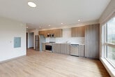 Located in Burlington, WA, this efficiently designed single-floor unit offers a thoughtful open-concept layout that combines the kitchen and living area into one cohesive space. Light-toned hardwood-style flooring runs throughout, creating a clean, modern aesthetic that enhances the sense of openness.

The kitchen is well-appointed with warm wood-grain cabinetry extending from floor to ceiling, providing ample storage. Stainless steel appliances include a refrigerator, range with oven, microwave, and dishwasher, all complemented by light-colored countertops and a white tile backsplash. The adjoining living area benefits from a pair of large windows that frame views of surrounding greenery and the Burlington streetscape, flooding the space with natural light.

The bathroom is clean and functional, finished with the same light flooring found throughout the unit. It includes a full soaking tub with shower, a wood-accented vanity, and a stacked washer and dryer unit — a practical convenience tucked neatly within the space. A closet near the entry provides additional storage.

Recessed lighting throughout and a neutral white-and-warm-wood palette give the unit a fresh, contemporary feel, making efficient use of every square foot in a well-organized, modern residential setting.