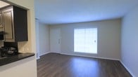 This single-floor space presents a straightforward and functional layout designed for comfortable everyday living. The living area serves as the focal point of the space, featuring rich, dark hardwood floors that extend throughout and provide a warm, grounding contrast to the light neutral-toned walls. A large window dressed with horizontal blinds allows an abundance of natural light to fill the room, creating a bright and welcoming atmosphere. A solid white entry door with brushed hardware offers a clean, classic entryway into the space. The walls are painted in a soft, understated neutral tone that complements the flooring and contributes to the overall sense of openness. The textured ceiling adds a subtle dimension to the interior finish. White baseboards line the perimeter of the room, providing a crisp, polished edge to the overall aesthetic. The open feel of the living area, combined with the natural light and durable flooring, makes this a practical and inviting space. The clean, minimal design ensures versatility, allowing the space to be easily personalized to suit a variety of interior styles and furnishing preferences.