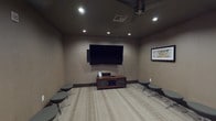 This well-appointed game room is designed for both entertainment and social gatherings, offering a cohesive layout across a single floor. The space is divided into distinct yet connected zones, each serving a unique purpose while maintaining a unified, sophisticated aesthetic throughout.

The media lounge area anchors one section of the room, where a large wall-mounted flat-screen television takes center stage above a wood media console. Recessed ceiling lighting and a ceiling fan provide both ambiance and comfort, while an array of modern round ottomets on swivel bases offer flexible, casual seating for multiple guests. The light-toned striped carpeting adds texture and warmth underfoot.

An adjacent seating nook features a generous sectional sofa in a plush gray upholstery, paired with a dark coffee table — an inviting retreat for relaxed conversation or gaming. Decorative wall art and sculptural wall accents add personality to the space.

A separate card and game table room, accessible through an open archway, is appointed with a round gaming table surrounded by upholstered chairs and illuminated by an elegant pendant light. Framed artwork lines the walls, reinforcing the room's refined character. A transitional wingback chair positioned near the entryway provides a comfortable spot for spectators. Together, these spaces create an engaging, multi-use environment ideal for entertainment.