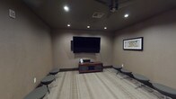 This well-appointed game room is designed for both entertainment and social gatherings, offering a cohesive layout across a single floor. The space is divided into distinct yet connected zones, each serving a unique purpose while maintaining a unified, sophisticated aesthetic throughout.

The media lounge area anchors one section of the room, where a large wall-mounted flat-screen television takes center stage above a wood media console. Recessed ceiling lighting and a ceiling fan provide both ambiance and comfort, while an array of modern round ottomets on swivel bases offer flexible, casual seating for multiple guests. The light-toned striped carpeting adds texture and warmth underfoot.

An adjacent seating nook features a generous sectional sofa in a plush gray upholstery, paired with a dark coffee table — an inviting retreat for relaxed conversation or gaming. Decorative wall art and sculptural wall accents add personality to the space.

A separate card and game table room, accessible through an open archway, is appointed with a round gaming table surrounded by upholstered chairs and illuminated by an elegant pendant light. Framed artwork lines the walls, reinforcing the room's refined character. A transitional wingback chair positioned near the entryway provides a comfortable spot for spectators. Together, these spaces create an engaging, multi-use environment ideal for entertainment.