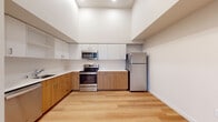 This thoughtfully designed single-floor unit offers a clean, contemporary aesthetic throughout. The open-concept living and kitchen area is anchored by soaring ceilings that lend an airy, expansive feel to the space. Light hardwood flooring runs seamlessly through the main areas, complementing the neutral white walls and creating a bright, welcoming atmosphere.

The kitchen is a standout feature, showcasing a two-tone cabinetry design with warm wood lower cabinets and crisp white upper cabinets extending nearly to the ceiling for generous storage. A full suite of stainless steel appliances — including a refrigerator, electric range, over-the-range microwave, and dishwasher — is set against clean white countertops, delivering a modern and functional workspace.

A hallway connects the main living area to the private quarters of the unit. The bedroom is generously proportioned with soaring ceilings, a wall-mounted climate control unit, and a window that frames views of the surrounding neighborhood, filling the room with natural light. The bedroom is finished with plush carpeting, adding warmth and comfort. An adjacent walk-in closet provides ample storage with wire shelving along the walls.

The full bathroom is finished consistently with the rest of the unit, featuring a combined tub and shower, a vanity with wood cabinetry, and light hardwood flooring.