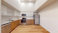 This thoughtfully designed single-floor unit offers a clean, contemporary aesthetic throughout. The open-concept living and kitchen area is anchored by soaring ceilings that lend an airy, expansive feel to the space. Light hardwood flooring runs seamlessly through the main areas, complementing the neutral white walls and creating a bright, welcoming atmosphere.

The kitchen is a standout feature, showcasing a two-tone cabinetry design with warm wood lower cabinets and crisp white upper cabinets extending nearly to the ceiling for generous storage. A full suite of stainless steel appliances — including a refrigerator, electric range, over-the-range microwave, and dishwasher — is set against clean white countertops, delivering a modern and functional workspace.

A hallway connects the main living area to the private quarters of the unit. The bedroom is generously proportioned with soaring ceilings, a wall-mounted climate control unit, and a window that frames views of the surrounding neighborhood, filling the room with natural light. The bedroom is finished with plush carpeting, adding warmth and comfort. An adjacent walk-in closet provides ample storage with wire shelving along the walls.

The full bathroom is finished consistently with the rest of the unit, featuring a combined tub and shower, a vanity with wood cabinetry, and light hardwood flooring.