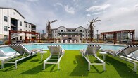 This outdoor amenity space is the centerpiece of a modern residential community, designed to deliver a resort-style experience for its residents. At the heart of the space is a generously sized swimming pool with crystal-clear blue water, framed by a wide, tiled pool deck that provides ample room for lounging and circulation. The pool's sweeping curved shape adds a dynamic visual element to the overall layout.

Surrounding the pool, multiple seating zones offer a variety of relaxation options. Lush artificial turf areas are furnished with contemporary striped chaise lounges and side tables, creating vibrant green pockets that contrast beautifully with the neutral tones of the hardscape. Additional lounge chairs line the tiled perimeter, offering both sun and shade options throughout the day.

Two rustic wood pergolas anchor opposite ends of the deck, providing shaded gathering points with a warm, inviting aesthetic. A covered cabana area at the far end of the pool features a ceiling fan, lounge seating, and what appears to be an outdoor screen — ideal for community events or casual outdoor entertainment.

Tall palm trees punctuate the space, reinforcing the resort ambiance, while thoughtfully landscaped planting beds with native shrubs and ground cover add texture and greenery throughout. The entrance to the amenity area is marked by a striking limestone-clad building facade with large glass doors, black metal fencing, and a stone-paved walkway — creating a polished and welcoming arrival experience.