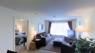 This single-floor unit offers a well-arranged living space centered around a large window dressed with deep burgundy curtains, allowing ample natural light to brighten the room. The living area features plush gray sofas arranged around a patterned area rug, with a flat-screen television positioned on a decorative media console along the opposite wall. A table lamp on a dark wood end table adds warm ambient lighting, complementing the neutral-toned carpet and light-colored walls throughout. Decorative mirror panels mounted on the wall provide an artistic accent to the space. An open entryway connects the living area directly to a bedroom, which is partially visible and contains a neatly made bed with patterned bedding and a bedside lamp, maintaining the warm and comfortable atmosphere found throughout the unit. The ceiling features a centrally mounted light fixture, and a smoke detector is also present, noting practical safety considerations. The overall ambiance is cozy and inviting, with a cohesive neutral color palette accented by warm lighting and thoughtful furnishings that maximize both comfort and functionality.