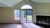 This single-floor space makes a striking first impression with its dramatic vaulted ceilings that rise to a pronounced peak, creating an expansive, airy atmosphere throughout the main living area. A standout architectural feature is the large arched transom window positioned high on the wall, which floods the room with abundant natural light and adds a touch of elegant design to the space.

Floor-to-ceiling sliding glass doors open directly onto a private outdoor balcony or patio, seamlessly connecting the interior living area to the outside. A secondary side window further enhances the natural brightness of the room, offering additional views of the surrounding greenery.

The living area is anchored by a classic fireplace with a clean white mantel and dark stone surround, serving as a refined focal point. The neutral-toned walls and light carpeted flooring complement the overall warm and inviting ambiance of the space.

The combination of vaulted ceilings, generous natural light from multiple windows, and direct outdoor access gives this space a sense of openness and volume that extends well beyond its physical footprint. The architectural details, including the arched window and fireplace, lend the space a timeless character suited for comfortable, everyday living.
