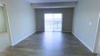 This single-floor space offers a clean, modern aesthetic defined by thoughtful design details and an inviting sense of openness. The main living area features smooth hardwood-style flooring in a warm, medium-toned finish that reflects natural light beautifully, enhancing the overall brightness of the room. Neutral gray walls provide a versatile backdrop, complementing the crisp white baseboards and ceiling trim that frame the space with a polished look. A prominent sliding glass door serves as the focal point of the living area, flooding the interior with abundant natural light and providing seamless access to a private balcony enclosed by a classic metal railing. The outdoor views visible through the glass add a sense of depth and connection to the surrounding environment. Subtle architectural details, including a structural ceiling beam, add character without compromising the clean lines of the interior. The layout feels open and airy, lending itself well to a variety of furniture arrangements. The overall ambiance is calm, bright, and contemporary, making it a highly functional and visually appealing space suited for comfortable everyday living.