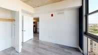 Located in the heart of Miami's vibrant Brickell neighborhood, this single-floor unit offers a compelling blend of industrial and contemporary design elements. The open living area is defined by striking floor-to-ceiling windows that frame expansive panoramic views of the Miami skyline and surrounding cityscape, flooding the space with abundant natural light throughout the day. Raw exposed concrete ceilings add an industrial character that contrasts beautifully with the smooth, light-toned wood-look flooring found throughout. Bold navy-blue accent walls provide a dramatic focal point in the main living area, enhancing the modern aesthetic. The sleek, galley-style kitchen is outfitted with stainless steel appliances, contemporary gray cabinetry, a clean white countertop, and a under-mount sink with a modern pull-down faucet — functional and stylish in equal measure. The bathroom features large-format tile surrounds in a warm neutral tone, a soaking tub, and a clean white vanity, offering a clean and practical finish. Additional rooms continue the cohesive design language with the same flooring, neutral white walls, and exposed ceilings, maintaining a consistent modern feel throughout the entire unit.