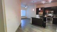 This single-story space features an open-concept design that seamlessly connects the kitchen and living areas, creating a cohesive and functional layout. The flooring throughout is finished in hardwood-style planks in a warm neutral tone, lending a clean and contemporary feel to the interior.

The kitchen is appointed with dark espresso-toned cabinetry that extends to the upper cabinets, providing ample storage. A matching dark-finished breakfast bar serves as a natural divider between the kitchen and living space, and two striking red pendant lights hang above it, adding a bold design accent. Stainless steel appliances complete the modern kitchen aesthetic.

The living area is well-lit by a window that allows natural light to fill the room, complemented by a ceiling fan with an integrated light fixture. The neutral beige walls provide a versatile backdrop throughout the space.

Overall, the unit presents a modern and practical layout with consistent design elements, balancing function and style through its contrasting dark fixtures against light walls and flooring.