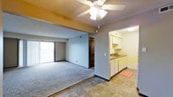 This single-floor space offers a practical and functional layout designed for comfortable everyday living. The main living area is carpeted and bathed in natural light from a sliding glass door that opens to an outdoor balcony, providing pleasant views of the surrounding area. A ceiling fan with an integrated light fixture is centrally mounted overhead, ensuring year-round comfort and air circulation. Neutral-toned walls in a cool gray hue lend a calm, understated ambiance throughout the space. A closet with louvered bifold doors is conveniently positioned within the living area, offering ample storage. The kitchen is directly adjacent to the living space, defined by tile flooring and a seamless transition between the two areas. It features white cabinetry above and below the counter, a sink with a window view, and generous counter space suitable for everyday meal preparation. Under-counter appliances are integrated neatly into the cabinetry. The kitchen's bright, well-lit environment is enhanced by overhead lighting. Together, the open flow between the living and kitchen areas creates a cohesive, easy-to-navigate layout that maximizes the utility of the available space.