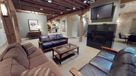 This distinctive space masterfully blends historic industrial character with modern comfort. Throughout, exposed heavy timber beams and structural columns form the architectural backbone of the interior, creating a warm, loft-like atmosphere with notable ceiling height. The original brick walls, visible in the dining and lounge areas, add authentic texture and visual depth that anchors the space in its historic roots.

The main lounge area is anchored by a sleek, wall-mounted fireplace flanked by flat-screen televisions, surrounded by generous leather seating and a wood-and-glass coffee table — ideal for relaxed gatherings. Adjacent to the lounge, a dedicated dining area features multiple high-top round and rectangular tables with solid wood chairs, all set against a backdrop of exposed brick and windows dressed with warm wood blinds. Large-format tile flooring grounds this section of the space.

The fully equipped kitchen is a standout feature, offering rich knotty wood cabinetry, dark granite countertops, a mosaic tile backsplash, and stainless steel appliances including a refrigerator, stove, microwave, and dishwasher. A central island with additional counter space enhances functionality. Recessed lighting and pendant fixtures throughout provide warm, even illumination, completing the inviting, versatile character of this well-appointed space.