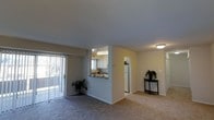 This single-floor residence offers an open and functional layout designed for comfortable everyday living. The main living area is finished with wall-to-wall carpeting in a neutral tone, creating a warm and cohesive atmosphere. A sliding glass door fitted with vertical blinds lines one wall, allowing natural light to filter into the space while providing access to an outdoor area.

The kitchen is positioned adjacent to the living area and is accessible via a pass-through window with a countertop overhang, ideal for casual dining or entertaining. The kitchen features white cabinetry, a white refrigerator, and a window above the sink that brings in additional natural light. Overhead lighting illuminates the kitchen effectively.

Neutral beige walls run throughout the space, maintaining a clean and consistent aesthetic. A hallway visible from the living area leads to additional rooms, hinting at a well-organized floor plan. Decorative touches, including potted plants and a small accent table, help convey the livable scale of the space. Overall, the unit presents a practical and inviting environment with a straightforward, single-level design.