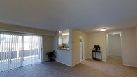 This single-floor residence offers an open and functional layout designed for comfortable everyday living. The main living area is finished with wall-to-wall carpeting in a neutral tone, creating a warm and cohesive atmosphere. A sliding glass door fitted with vertical blinds lines one wall, allowing natural light to filter into the space while providing access to an outdoor area.

The kitchen is positioned adjacent to the living area and is accessible via a pass-through window with a countertop overhang, ideal for casual dining or entertaining. The kitchen features white cabinetry, a white refrigerator, and a window above the sink that brings in additional natural light. Overhead lighting illuminates the kitchen effectively.

Neutral beige walls run throughout the space, maintaining a clean and consistent aesthetic. A hallway visible from the living area leads to additional rooms, hinting at a well-organized floor plan. Decorative touches, including potted plants and a small accent table, help convey the livable scale of the space. Overall, the unit presents a practical and inviting environment with a straightforward, single-level design.