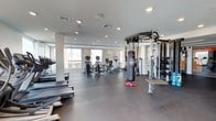 This fitness center offers a spacious, open-plan layout designed to accommodate a wide range of workout styles and fitness goals. The interior features a clean, contemporary aesthetic with neutral tones, recessed lighting, and large windows that flood the space with natural light and views of the surrounding area.

The cardio zone is well-appointed with a selection of treadmills and elliptical machines, providing ample space between each unit for a comfortable workout experience. A multi-functional cable and pull-up station anchors the strength training area, complemented by a full rack of dumbbells in graduated weights, catering to both beginners and advanced users. A heavy-duty punching bag adds a dynamic element for boxing and functional fitness training.

The flooring throughout is a durable, dark rubberized surface, ideal for high-impact activities and easy maintenance. Structural columns are cleanly integrated into the design without interrupting the flow of the space. The ceiling height contributes to an airy, open atmosphere, while the recessed lighting ensures consistent illumination across all workout zones. Overall, this fitness center is thoughtfully designed to deliver a comprehensive and energizing exercise environment.