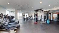 This fitness center offers a spacious, open-plan layout designed to accommodate a wide range of workout styles and fitness goals. The interior features a clean, contemporary aesthetic with neutral tones, recessed lighting, and large windows that flood the space with natural light and views of the surrounding area.

The cardio zone is well-appointed with a selection of treadmills and elliptical machines, providing ample space between each unit for a comfortable workout experience. A multi-functional cable and pull-up station anchors the strength training area, complemented by a full rack of dumbbells in graduated weights, catering to both beginners and advanced users. A heavy-duty punching bag adds a dynamic element for boxing and functional fitness training.

The flooring throughout is a durable, dark rubberized surface, ideal for high-impact activities and easy maintenance. Structural columns are cleanly integrated into the design without interrupting the flow of the space. The ceiling height contributes to an airy, open atmosphere, while the recessed lighting ensures consistent illumination across all workout zones. Overall, this fitness center is thoughtfully designed to deliver a comprehensive and energizing exercise environment.