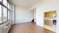 This thoughtfully designed residence offers a functional and open layout spread across a single floor, with warm hardwood flooring flowing throughout the main living areas and bedrooms. The spacious living room features large windows that invite generous natural light and frame elevated city views, while a staircase leads to an additional upper level, adding vertical dimension to the space.

The kitchen is well-equipped with light wood cabinetry extending to the ceiling, white countertops, and a full suite of stainless steel appliances including a gas range, built-in microwave, and dishwasher. The clean, modern design of the kitchen provides ample storage and prep space.

The bedrooms are bright and airy, each benefiting from wide windows with urban vistas and radiator heating units beneath the sills. The crisp white walls create a neutral, versatile backdrop throughout.

Two full bathrooms are finished in a clean, all-white palette, each featuring a bathtub with tiled surround, a vanity with sink, and a large mirror. A dedicated laundry area with washer hookups and overhead shelving adds everyday convenience. Overall, the space presents a clean, modern aesthetic with practical amenities throughout.