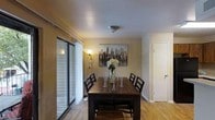 This single-floor space features a welcoming dining area that serves as a central gathering point. The room is finished with warm hardwood flooring that adds a natural, inviting quality to the space. Neutral beige walls provide a versatile backdrop, complemented by a flush-mount ceiling light that offers soft, even illumination throughout the room. A large sliding glass door along one wall floods the dining area with natural light while offering a view of the exterior brick surroundings, and is dressed with vertical blinds for adjustable privacy. The dining space comfortably accommodates a dark wood rectangular table paired with matching ladder-back chairs, making it well-suited for everyday meals or gatherings. Decorative touches, including a cityscape canvas print on the wall and a contemporary floor lamp in the corner, contribute to the room's modern yet cozy ambiance. A doorway visible at the far end of the room connects to additional areas of the space, suggesting a functional and straightforward layout throughout the single-level floor plan.