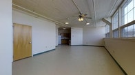 This single-floor space offers a generous, open-concept layout defined by its industrial-influenced architectural character. The expansive main room immediately draws attention with its notably high ceilings finished in painted wood planking, complemented by exposed structural elements and ceiling fans that enhance airflow and add to the loft-like atmosphere. Large windows line the upper portion of the walls, flooding the interior with abundant natural light and contributing to an airy, open ambiance. The neutral-toned walls and concrete-style flooring provide a clean, versatile canvas throughout the main living area. Toward one end of the space, a compact kitchen is seamlessly integrated, featuring warm wood cabinetry, a white refrigerator, and a range, all set against a functional layout that makes efficient use of the available footprint. Adjacent to the kitchen, a bathroom area with essential fixtures is also present. The overall design reflects a utilitarian yet adaptable character, well-suited for a variety of uses. The combination of open floor space, elevated ceilings, plentiful natural light, and practical amenities makes this a distinctive and functional space with strong potential for both residential and creative applications.