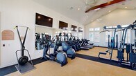 This fitness center offers a bright, open workout environment defined by its impressive vaulted ceilings with exposed dark wood beam accents and multiple ceiling fans that promote airflow throughout the space. The architectural design creates a generous, airy atmosphere that enhances the overall workout experience.

The cardio zone is outfitted with a full row of treadmills, elliptical trainers, and additional cardio machines, all positioned to take advantage of the natural light streaming through the numerous large windows that line the walls. Large wall-mounted mirrors and flat-screen televisions are strategically placed throughout, providing both visual expansion of the space and entertainment options during workouts.

The strength training area features a comprehensive array of equipment, including multi-station cable machines, leg press and extension machines, a dumbbell rack with an adjustable bench, and various selectorized machines. This section benefits equally from the soaring ceiling height and abundant window placement.

The flooring combines neutral carpet tiles in the open areas with darker rubberized flooring beneath the equipment zones. French doors on one side open to an outdoor area, allowing natural ventilation and easy access. The overall ambiance is clean, modern, and well-lit, creating a welcoming and functional environment for a full range of fitness activities.