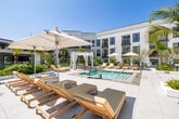Located at 3988 E Hardy Street in Inglewood, California, this expansive outdoor amenity space is designed to evoke a resort-like atmosphere in the heart of Southern California. The centerpiece of the space is a rectangular pool surrounded by wide-format stone pavers that provide ample room for circulation and relaxation. Teak wood chaise lounges are arranged in clusters around the pool deck, complemented by large white market umbrellas offering shade throughout the day.

Private cabanas with white draped curtains and striped interior cushions are positioned at key points around the perimeter, providing semi-enclosed retreats for residents. Comfortable seating vignettes—featuring cushioned sofas, teak coffee tables, and wrought-iron accent chairs—are thoughtfully distributed across the courtyard, creating distinct social and lounge zones.

The landscaping is a standout feature, with mature palm trees, tropical banana-leaf plants, ornamental grasses, and lush greenery integrated throughout the space. Raised planting beds with glass railings add dimension to the layout and define pathways between areas. A pergola-covered entry point with bold red doors and glass walls anchors one end of the courtyard, adding architectural contrast to the crisp white building façade that frames the entire space on multiple sides.