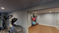 The Mansions fitness center is a thoughtfully designed, single-floor workout facility that provides residents with a comprehensive exercise environment. The space is finished with cool grey walls, white crown molding, and a combination of durable wood-look flooring and low-pile carpeting, creating a clean and functional aesthetic throughout.

The main workout area is equipped with a range of cardio machines, including an elliptical trainer, a recumbent bike, and an upright cycle, all positioned to allow comfortable use. A dedicated stretch and functional training alcove features a vertical exercise ball rack stocked with multiple stability balls in varying sizes, alongside a set of dumbbells — ideal for flexibility and strength training routines.

Recessed lighting illuminates the space evenly, while a wall-mounted flat-screen television near the entrance provides entertainment during workouts. The facility is accessed through a set of double glass doors that allow natural light to filter into the entry area, which also includes a welcome mat and a small shelving unit for personal belongings.

Additional doors within the space lead to separate utility or storage rooms, adding to the facility's practicality. The overall layout is open and well-organized, making it easy for users to move between equipment and exercise zones with ease.