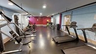 This fitness center is a dedicated workout space designed to accommodate a wide range of exercise needs. The room features durable rubber tile flooring, which provides both safety and comfort underfoot during training sessions. Recessed lighting illuminates the space evenly from above, ensuring a bright and functional environment throughout.

The cardio section is outfitted with multiple treadmills and stationary bikes, positioned to make efficient use of the available floor space. On the strength training side, the facility includes a weight bench, a cable or resistance machine, a dumbbell rack, and a selection of free weights, offering users ample options for resistance-based workouts. Stability balls and additional accessories are also present, supporting flexibility and core training.

The walls are painted in a bold deep red accent color, contrasted against lighter painted concrete block walls, giving the space an energetic and motivating atmosphere. Large mirrors line one wall, a practical feature that allows users to monitor their form during exercise. The space is accessed through a solid entry door and is well-organized, with equipment arranged to maximize open floor area for stretching or floor exercises. Overall, this fitness center provides a comprehensive and functional environment suited to a variety of fitness routines.