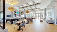 Located at 12609 Matisse Cir in New Port Richey, Florida, this single-story community clubhouse showcases a thoughtfully designed coastal aesthetic that blends contemporary architecture with warm, inviting interiors. The space is organized into several distinct yet interconnected zones that flow seamlessly from one to the next across light-toned hardwood-style flooring.

The entry foyer sets an impressive tone with an illuminated reception desk, dramatic blue accent walls, shiplap detailing, and sculptural coastal artwork. A bold spherical chandelier anchors the space overhead. Adjacent lounge areas feature soaring ceilings with coffered and tray detailing, large-format windows, and glass-paned French doors that flood the interior with natural light and frame views of a sunny outdoor pool area.

Social seating zones offer a mix of plush sofas, lounge chairs, and casual dining tables with modern pendant lighting overhead. A dedicated lounge nook with bold blue walls and a chevron-patterned barn door provides a more intimate retreat. The well-appointed community kitchen features an expansive white quartz island with wicker bar seating, stainless steel appliances, open shelving with décor accents, subway tile backsplash, and mounted televisions. Throughout, coastal-inspired artwork, lush indoor plants, and carefully curated furnishings contribute to a bright, welcoming ambiance.
