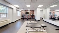 Located at 23 Spring St in Hamilton, Ontario, this fitness room is a compact yet thoughtfully designed exercise space that offers a comprehensive range of workout options. The room features a clean, modern aesthetic with white walls, a drop-panel ceiling equipped with recessed LED lighting, and durable rubber-and-speckled flooring that transitions between darker and lighter zones to define different workout areas.

A striking feature wall adorned with bold, colorful graphic mural wallpaper anchors one end of the room, providing a visually energizing backdrop for cardio equipment including treadmills and a stationary bike. A flat-screen television is mounted on this accent wall, adding entertainment for users during workouts.

The strength training area is well-appointed with a multi-tier dumbbell rack stocked with a full range of hex weights, adjustable workout benches, and an assortment of stability balls and resistance accessories neatly organized on a dedicated storage rack. Full-length mirrors line one wall, enhancing the sense of space and allowing users to monitor their form.

Natural light enters through windows along the perimeter, offering views of the surrounding neighborhood. A wall-mounted HVAC unit ensures year-round climate comfort. The space is practical and functional, with cubbies for storage and a fire extinguisher for safety.