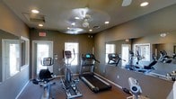 This fitness center is thoughtfully designed to provide a comprehensive workout environment within a single, open-plan room. The space is furnished with a variety of cardio equipment, including a treadmill, elliptical trainer, and stationary bike, offering users multiple options for aerobic exercise. A set of free weights is also available, catering to strength training needs. The room features neutral-toned walls that create a calm, focused atmosphere, while recessed lighting and a ceiling fan ensure the space remains well-lit and comfortably ventilated during workouts. A large wall mirror spans one side of the room, enhancing the sense of space and allowing users to monitor their form. Multiple windows allow natural light to filter in, contributing to an open and energizing ambiance. Emergency exit signage is clearly visible, reflecting a commitment to safety. The durable flooring is suited for high-traffic fitness use. Whether for a quick cardio session or a full strength-training routine, this fitness center is equipped to meet a variety of exercise needs in a practical and welcoming environment.