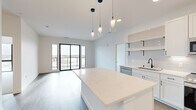This well-appointed 2-bedroom, 2-bathroom unit offers a thoughtfully designed single-level layout with a clean, contemporary aesthetic throughout. The open-concept kitchen and living area serves as the heart of the unit, anchored by a large quartz-topped island with striking veining. The kitchen is outfitted with crisp white shaker cabinets accented by matte black hardware, open shelving, a subway tile backsplash, and a full suite of stainless steel appliances including a range, over-the-range microwave, and refrigerator. Pendant lighting with mixed glass fixtures adds a modern, artistic touch above the island. The living area extends toward floor-to-ceiling black-framed sliding glass doors that open to a private balcony, flooding the space with natural light and offering open views of the surrounding landscape. Bedrooms are carpeted and bright, each featuring large black-framed windows with expansive outdoor views. The spacious primary bedroom includes a generously sized walk-in closet with built-in rod systems. Both bathrooms are finished with large-format tile flooring, white cabinetry with matte black fixtures, quartz vanity tops, and well-lit mirrors. A dedicated laundry room houses a stacked washer and dryer unit. Wood-look flooring runs throughout the main living areas, tying together this modern, move-in-ready space.
