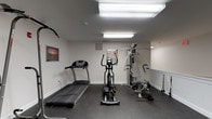 This fitness center is a dedicated, single-level exercise space designed to support a comprehensive workout routine. The room features durable rubber flooring throughout, providing both safety and comfort underfoot during exercise. Overhead fluorescent lighting illuminates the space evenly, ensuring excellent visibility across the entire room.

The cardio section is well-appointed with a True treadmill and an elliptical trainer, offering users a variety of low-impact and high-intensity aerobic options. A stationary recumbent bike adds another cardio alternative on the opposite side of the room. For strength training, the space includes a multi-function cable and weight machine, a freestanding pull-up and dip tower, and a dedicated dumbbell rack stocked with a selection of free weights.

A water cooler is conveniently positioned within the space, allowing users to stay hydrated during their workouts. Motivational framed artwork and informational posters are displayed on the neutral-toned walls, contributing to an energized atmosphere. The layout is thoughtfully organized to allow ample circulation between equipment. A separate adjacent area with tiled flooring provides additional open space, visible through a doorway, suggesting room for stretching or supplementary activities. Safety features, including a fire alarm pull station, are also present throughout the space.