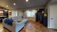 This single-floor space offers an open-concept layout that seamlessly connects the kitchen, dining, and living areas, creating a cohesive and functional environment. Rich hardwood-style flooring runs throughout, lending warmth and continuity to the entire space.

The kitchen is a standout feature, outfitted with warm-toned wood cabinetry, a decorative tile backsplash in a geometric pattern, and a full suite of stainless steel appliances including a French door refrigerator and dishwasher. A large central island with a granite countertop and industrial-style metal bar stools provides both additional workspace and casual seating. A rustic wood beam pendant light with mason jar fixtures hangs above the island, adding a charming, handcrafted accent. Navy-painted accent walls frame the kitchen area and contribute a bold design contrast.

The open living area is bright and welcoming, with recessed lighting and multiple windows allowing natural light to fill the space. A generously sized sectional sofa anchors the living room, complemented by a media cabinet and wall-mounted television. A secondary sitting area near the entrance features a cozy accent chair, wall décor, and double doors that suggest access to additional rooms or private offices. The overall ambiance is casual yet well-designed, blending rustic and modern elements throughout.
