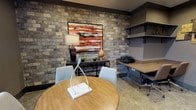 The business center at The Lodges On English Station is a thoughtfully designed single-floor workspace that balances functionality with refined aesthetics. A striking stacked stone accent wall serves as the room's focal point, anchoring a bold abstract artwork in warm reds and oranges that adds character to the space. Recessed lighting illuminates the room evenly, complementing the natural light that filters in through large windows dressed with sheer curtains, offering views of the surrounding landscape.

The workspace is organized to accommodate both individual and collaborative needs. A built-in countertop workstation with wall-mounted shelving runs along one side of the room, providing ample desk space for multiple users along with a printer for added convenience. Adjustable office chairs ensure comfortable, extended use. On the opposite end, a round wooden table with upholstered seating creates an informal meeting or collaboration area. Neutral-toned carpeting, dark crown molding, and taupe walls contribute to a cohesive, professional atmosphere throughout. Decorative wall art and accent pieces on open shelving add finishing touches that elevate the space beyond a purely utilitarian setting.