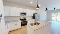 Located at 2700 Emerald Green Way in Knoxville, Tennessee, this thoughtfully designed one-bedroom, one-bathroom residence offers a clean, modern aesthetic throughout its well-utilized single floor. The open-concept kitchen and living area serves as the heart of the unit, featuring crisp white shaker cabinets extending to the ceiling, a white subway tile backsplash, light quartz countertops, and a central island with an undermount sink. Stainless steel appliances — including a range, microwave, and refrigerator — are complemented by stylish black pendant lighting and light wood-style flooring that flows throughout the main living spaces. The living area is bright and airy, equipped with a ceiling fan and a sliding glass door that opens to a private balcony with outdoor views. The bedroom offers plush carpeting, multiple windows that fill the room with natural light, and access to two closets, including a spacious walk-in closet with built-in wire shelving. The full bathroom is finished in a clean white palette with wood-style flooring, a combined tub and shower, and a vanity with black hardware accents. Additional conveniences include a dedicated laundry area, a utility room, and hallway spaces that connect the layout efficiently.
