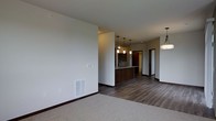 Located at 310 Samuel Dr in Madison, Wisconsin, this single-floor residence offers a thoughtfully designed open-concept layout that seamlessly connects the living and kitchen areas. The living space features plush carpeting, crisp white walls, and dark wood baseboards that provide a clean, modern contrast throughout. A floor-mounted vent ensures year-round climate comfort, while a wall-mounted thermostat adds convenience.

The kitchen is a standout feature, anchored by a rich, dark wood-toned peninsula that doubles as a breakfast bar. Sleek pendant lighting hangs above the peninsula, complementing a matching chandelier-style fixture in the adjacent dining area, creating a warm and inviting atmosphere. The kitchen and connecting hallway are finished with durable hardwood-style flooring that extends toward the rear of the unit.

Dark cabinetry lines the kitchen, contributing to the cohesive, contemporary aesthetic carried throughout the space. A hallway beyond the kitchen leads to additional rooms, offering a practical and well-organized floor plan. The neutral color palette, combination of carpet and hard-surface flooring, and carefully chosen lighting fixtures give this space a polished, move-in-ready feel well-suited for modern living in the Madison area.