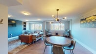 This thoughtfully designed single-floor space blends modern aesthetics with functional layout, offering a seamlessly connected living and dining environment. The open-concept main area is anchored by warm hardwood flooring that extends throughout, complemented by soft blue-gray walls that create a calm, cohesive atmosphere.

The living area is divided into distinct zones, featuring a media wall with a bold teal accent, built-in shelving, and a wall-mounted television. A tufted leather sofa, plush accent chairs, and a round ottoman coffee table furnish this space, while large windows frame views of mature, wooded surroundings, filling the room with natural light.

A round dining table with metal-accented chairs occupies a central position in the open area, beneath a striking sputnik chandelier that serves as an architectural focal point. Multiple seating arrangements with wingback chairs create additional conversational areas near the windows.

A built-in wet bar with rich mahogany cabinetry and dark countertops connects to an adjacent room accessible through a sleek glass sliding door. Clean white walls, flush-mount lighting, and generous baseboard trim throughout contribute to the space's polished, contemporary character.