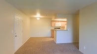 This single-floor space features an open-concept layout that seamlessly connects the living area and kitchen. The living area is finished with wall-to-wall neutral-toned carpet, creating a warm and cohesive feel throughout the space. Smooth, light-colored walls and a flush ceiling light fixture contribute to a clean, understated aesthetic.

The kitchen is positioned at the rear of the open layout and is partially separated by a peninsula-style counter with a dark laminate top, providing a natural divide between the cooking and living areas while maintaining an open sightline. Light wood cabinetry lines the kitchen walls, offering ample storage space. Appliances include a built-in microwave and a range, both finished in white to complement the cabinetry. The kitchen's elevated counter section adds a casual dining or prep surface option.

Overall, the space maintains a simple, functional design with a neutral color palette of beige and white tones. The open floor plan maximizes the sense of space, making the interior feel bright and accessible. The layout is well-suited for everyday living with practical storage and cooking amenities integrated into the design.