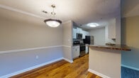Located at 100 Preston Woods Trail in Sandy Springs, GA, this one-bedroom, one-bathroom unit offers a well-organized single-floor layout designed for comfortable everyday living. The kitchen is a standout feature, configured in a classic galley style with white cabinetry, dark granite-style countertops, stainless steel appliances including a refrigerator, electric range, and microwave, and a dishwasher. A peninsula counter extends into the adjacent dining area, creating a natural transition between the two spaces. The dining area is finished with warm hardwood flooring and is highlighted by a pendant light fixture and crown molding detail. Hardwood flooring continues throughout the main living areas, lending a cohesive and polished look. The living room offers a comfortable, open feel with access to a private exterior door and windows that invite natural light. The bedroom is carpeted and generously sized, featuring a window with exterior views that bring in additional daylight. The bathroom includes a full tub-shower combination with white tile surround and hardwood flooring. A dedicated in-unit laundry room with a full-size washer and dryer adds significant everyday convenience. Neutral wall tones throughout create a clean, versatile backdrop suited to a variety of decor styles.