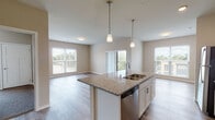 This well-appointed single-floor residence offers a thoughtfully designed open-concept layout that seamlessly connects the kitchen, dining, and living areas. The kitchen serves as the centerpiece of the main living space, featuring a large central island with a granite countertop, an undermount sink, and elegant pendant lighting overhead. White shaker-style cabinetry lines the walls alongside a full suite of stainless steel appliances, including a refrigerator, range, microwave, and dishwasher. Light wood-tone luxury vinyl plank flooring runs throughout the main living areas, contributing to a clean and contemporary aesthetic. The spacious living area is bathed in natural light, courtesy of large windows and a sliding glass door that leads to a private balcony with open outdoor views. The bedrooms are carpeted for comfort and feature generous windows that bring in natural light and views of the surrounding neighborhood. Two bathrooms are present, each finished with wood-look LVP flooring, clean white fixtures, and a minimalist design. Neutral warm beige tones are carried throughout the entire residence, creating a cohesive and inviting ambiance. The overall design balances functionality and modern style, making it a comfortable and practical living space.