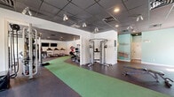 Located at 349 Campus Cove in Wilmington, NC, this fitness center offers a comprehensive workout environment spread across a generously sized single-floor facility. The space is divided into two interconnected exercise rooms, each designed to support a full range of fitness activities.

The main exercise area features durable rubber and green turf flooring, providing cushioning for both cardio and strength training. A row of treadmills lines one wall, accompanied by cable machines, weight benches, and a full dumbbell rack with wall-mounted mirrors for form monitoring. Flat-screen TVs are mounted for entertainment during workouts. The drop ceiling is fitted with recessed lighting and adjustable track lighting, ensuring a well-lit environment throughout.

The second exercise room is bathed in natural light from a bank of windows along one wall, offering views of the surrounding outdoors. Teal accent walls add a vibrant, energizing aesthetic to the space. This room is equipped with elliptical machines, a rowing machine, a power rack, kettlebells, medicine balls, stability balls, and TRX suspension training equipment, catering to functional and high-intensity training styles. A bold geometric wall mural adds a dynamic visual element.

A small amenity alcove with a water fountain and restroom access is conveniently located between the two rooms, completing this well-appointed fitness facility.