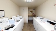 This shared laundry room is a single-story, self-contained facility designed to efficiently serve multiple users. The room is laid out with rows of top-loading washing machines and dryers positioned along both side walls, providing ample laundry capacity. The machines are white and uniformly arranged, creating an organized and functional environment.

The flooring consists of durable tile in a neutral tone, well-suited for the demands of a high-traffic utility space. The walls are painted in a light, cream-toned finish that keeps the room feeling bright and clean. Overhead fluorescent lighting ensures consistent illumination throughout, while two windows flanking the exterior door allow natural light to supplement the artificial lighting and provide views of the surrounding outdoor area.

At the rear of the room, a built-in countertop workspace offers a convenient folding and sorting surface. Above it, a bulletin board displays informational notices, and a fire extinguisher is mounted nearby for safety compliance. A posted sign near the entrance clearly indicates the laundry room's operating hours.

The exterior door provides direct outdoor access, contributing to natural ventilation. Overall, the space is practical, well-maintained, and thoughtfully equipped to accommodate the laundry needs of a shared residential setting.