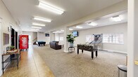 This generously sized single-floor community amenity space offers a well-appointed recreational environment designed for social engagement and entertainment. The open layout is anchored by a central corridor that seamlessly connects distinct activity zones, all finished with a combination of neutral carpet and tile flooring beneath bright, recessed fluorescent lighting.

The main game area is a standout feature, equipped with two full-size pool tables topped with vibrant blue felt, a foosball table, and a basketball arcade game — providing a variety of entertainment options for all types of guests. Multiple wall-mounted flat-screen TVs are positioned throughout the space for easy viewing from multiple vantage points.

A comfortable lounge area features leather sofas and a wooden coffee table, creating an inviting spot to relax between games. Structural columns are integrated throughout the space and contribute to the open, loft-like feel without obstructing sightlines. Natural light filters in through several windows along the perimeter walls, complementing the bright overhead lighting.

Near the entrance, a tiled foyer connects to a staircase and elevator, providing easy access and flow. Additional amenities include shelving units stocked with snacks and a vending machine, adding a convenient refreshment element. A potted indoor plant adds a touch of greenery to the otherwise clean, modern aesthetic.