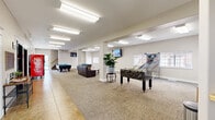 This generously sized single-floor community amenity space offers a well-appointed recreational environment designed for social engagement and entertainment. The open layout is anchored by a central corridor that seamlessly connects distinct activity zones, all finished with a combination of neutral carpet and tile flooring beneath bright, recessed fluorescent lighting.

The main game area is a standout feature, equipped with two full-size pool tables topped with vibrant blue felt, a foosball table, and a basketball arcade game — providing a variety of entertainment options for all types of guests. Multiple wall-mounted flat-screen TVs are positioned throughout the space for easy viewing from multiple vantage points.

A comfortable lounge area features leather sofas and a wooden coffee table, creating an inviting spot to relax between games. Structural columns are integrated throughout the space and contribute to the open, loft-like feel without obstructing sightlines. Natural light filters in through several windows along the perimeter walls, complementing the bright overhead lighting.

Near the entrance, a tiled foyer connects to a staircase and elevator, providing easy access and flow. Additional amenities include shelving units stocked with snacks and a vending machine, adding a convenient refreshment element. A potted indoor plant adds a touch of greenery to the otherwise clean, modern aesthetic.