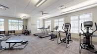 This fitness center is thoughtfully designed to support a comprehensive workout experience within a single-floor layout. The space is divided into distinct functional zones, each dedicated to a different training focus. The main cardio area features multiple treadmills and elliptical trainers, along with upright and stationary bikes, all positioned along a wall of large windows fitted with slatted blinds that allow generous natural light to fill the room. A ceiling fan overhead contributes to a comfortable workout environment, complemented by recessed and fluorescent lighting set within a drop-tile ceiling. A motivational wall decal reading "Everyday is a Good Day to Work Out" reinforces the energetic atmosphere. An adjacent strength training area houses a multi-station cable machine, a weight bench, and a full rack of dumbbells in graduated sizes, catering to a range of resistance training needs. A separate functional training zone provides medicine balls in various weights, resistance bands, a stability ball, and foam rollers stored in organized cubbies. Throughout, the flooring consists of durable patterned carpet tiles suited for high-traffic use. A wall-mounted television and a potted plant near the entrance add to the facility's welcoming and practical design. Large exterior-facing windows across multiple zones offer outdoor views and maintain a bright, open ambiance throughout.