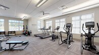 This fitness center is thoughtfully designed to support a comprehensive workout experience within a single-floor layout. The space is divided into distinct functional zones, each dedicated to a different training focus. The main cardio area features multiple treadmills and elliptical trainers, along with upright and stationary bikes, all positioned along a wall of large windows fitted with slatted blinds that allow generous natural light to fill the room. A ceiling fan overhead contributes to a comfortable workout environment, complemented by recessed and fluorescent lighting set within a drop-tile ceiling. A motivational wall decal reading "Everyday is a Good Day to Work Out" reinforces the energetic atmosphere. An adjacent strength training area houses a multi-station cable machine, a weight bench, and a full rack of dumbbells in graduated sizes, catering to a range of resistance training needs. A separate functional training zone provides medicine balls in various weights, resistance bands, a stability ball, and foam rollers stored in organized cubbies. Throughout, the flooring consists of durable patterned carpet tiles suited for high-traffic use. A wall-mounted television and a potted plant near the entrance add to the facility's welcoming and practical design. Large exterior-facing windows across multiple zones offer outdoor views and maintain a bright, open ambiance throughout.