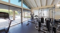 This fitness center offers a bright, open workout environment defined by striking vaulted ceilings with exposed white beams that lend a sense of spaciousness and architectural character. Large floor-to-ceiling sliding glass doors and expansive windows flood the space with natural light while providing direct views of a landscaped outdoor pool area, creating an inviting and energizing atmosphere for exercise.

The main workout floor is finished with durable rubber flooring throughout and is equipped with a comprehensive selection of fitness equipment, including a multi-station cable strength machine, a recumbent bike, an elliptical trainer, a treadmill, and a flat weight bench with free weights. A full-length mirror along one wall visually expands the space and supports proper form during exercise.

A built-in counter with warm wood cabinetry, a dark stone countertop, and a sink provides a convenient amenity station — ideal for towels, supplies, or quick refreshment. The warm yellow-toned walls complement the white ceiling structure, contributing to a clean and welcoming aesthetic. A private door offers access to an additional room or restroom. The single-floor layout ensures easy accessibility throughout, making this fitness center a functional and well-appointed space for a wide range of workout routines.