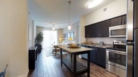 This single-floor residence features an open-concept layout that seamlessly connects the kitchen and living areas, creating a spacious and cohesive living environment. The kitchen is equipped with dark espresso-toned cabinetry that extends to the upper and lower sections, providing ample storage. Granite countertops add a touch of elegance, complemented by stainless steel appliances and a single-basin sink. A central island with a matching granite surface and lower shelving serves as both a functional workspace and a visual anchor for the space.

Wood-look tile flooring extends throughout, offering durability and a warm aesthetic. Pendant lighting above the island and a flush-mount ceiling fixture provide focused illumination in the kitchen zone. The living area transitions naturally from the kitchen, featuring a ceiling fan with integrated lighting. A sliding glass door at the far end of the living space allows natural light to filter through sheer curtains, brightening the interior. Neutral wall tones and a clean, contemporary design palette lend the space a modern yet welcoming ambiance.